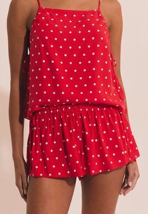 Mujer con top sin mangas rojo y pantalones cortos a juego con lunares blancos, de pie con los brazos relajados a los lados.