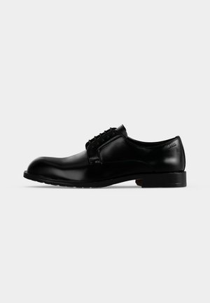 Scarpa elegante da uomo in pelle nera lucida con lacci, tacco basso e punta leggermente arrotondata, mostrata di lato su uno sfondo semplice.