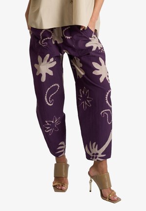 Pantaloni larghi viola con grandi motivi floreali e paisley beige, indossati con sandali beige con tacco alto e punta aperta, modello in piedi con le mani nelle tasche.