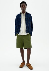 Denim jacket with front pockets, light striped shirt, olive green shorts, and brown moccasin-style shoes. Neutral background.