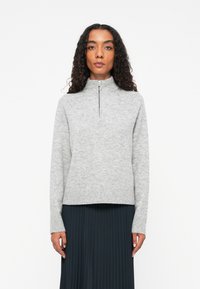 Suéter gris de cuello alto con cierre de media cremallera. Material de punto suave, puños y dobladillo acanalados, combinado con una falda oscura plisada.