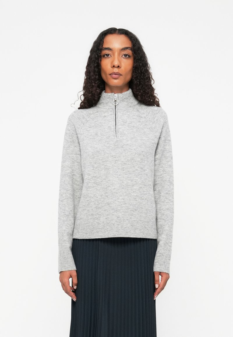 Maglione grigio a collo alto con chiusura a zip a un quarto. Materiale a maglia morbido, con polsini e orlo a coste, abbinato a una gonna scura plissettata.