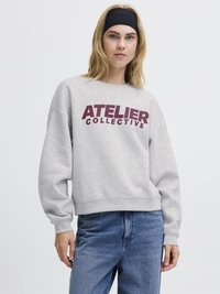 Sivý sveter s nápisom "ATELIER COLLECTIVE" v burgundskej farbe. Má voľný strih, okrúhly výstrih a manžety na rukávoch. Kombinovaný s modrými džínsami.
