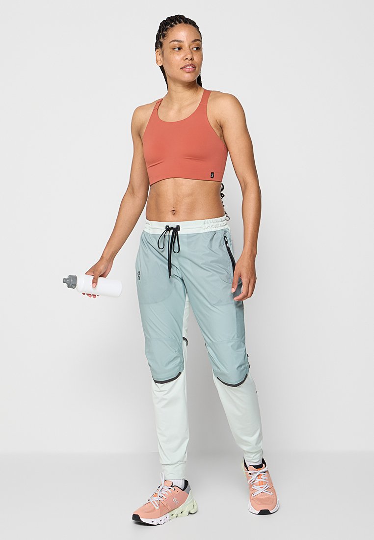 Leichtes Sportoutfit, bestehend aus einem rostfarbenen Sport-BH und aquafarbenen Laufhosen mit schwarzen Akzenten; kombiniert mit einer Wasserflasche und Laufschuhen.