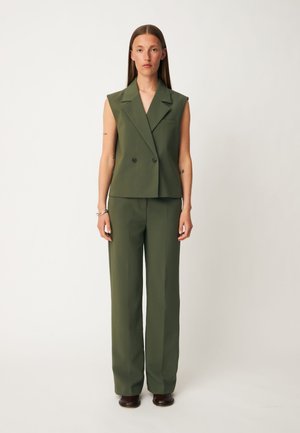 Femme debout portant un blazer sans manches vert olive et un pantalon large assorti avec des chaussures marron sur un fond uni.