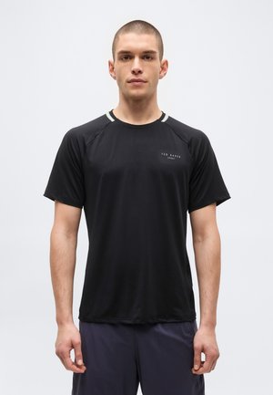 Jaunas vyras, žvelgiantis į priekį, dėvi juodą trumpomis rankovėmis sportinį marškinėlį su "Ted Baker Sport" logotipu ant šviesios fono.