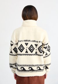 Krémový fleecový pulovr s černými geometrickými vzory, vysokým límcem a dlouhými rukávy. Texturovaná látka s útulným, měkkým vzhledem.