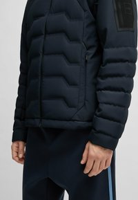 Marinblå puffer-jacka med en kviltad design, som har dragkedjestängning, sidofickor och en strukturerad svart detalj på axeln.