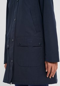 Veste bleu marine avec poches à bouton-pression, fermeture éclair visible et logo "Barbour" brodé sur le rabat de la poche gauche, portée par une personne.