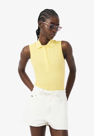 Vrouw met gevlochten haar die roze zonnebril draagt, een mouwloos geel poloshirt en witte shorts met handen in de zakken.