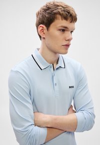Ανοιχτό μπλε πόλο με μακριά μανίκια και γιακά με μαύρες και λευκές ρίγες. Φέρει το λογότυπο της μάρκας "BOSS" σε μαύρο στο στήθος.