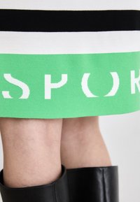 Čierno-biela pletená sukňa s odvážnym zeleným pásom na spodnom leme, s bielymi blokovými písmenami, ktoré hlásajú "SPOK." Hladká textúra a priliehavý strih.