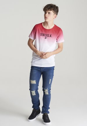 Czerwono-biała koszulka z gradientem z logo "SIKSILK", zestawiona z przetartymi niebieskimi dżinsami i czarnymi trampkami na białej podeszwie.