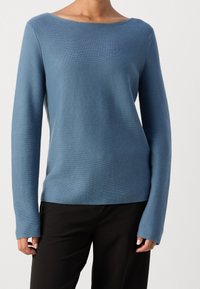 Maglione blu lavorato a maglia con un ampio scollo e maniche lunghe, caratterizzato da una vestibilità leggermente morbida e una texture a coste. Un paio di pantaloni neri è visibile.