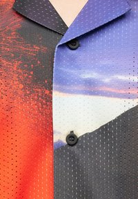 Chemise à boutons avec un motif en tissu perforé, présentant un dégradé en rouge, noir et un ciel bleu avec des nuages.
