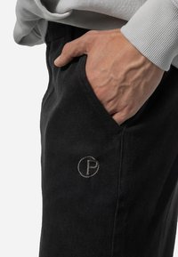 Black pants with a smooth texture, featuring a side pocket and an embroidered circular logo in gray. Close-up view with hand visible.