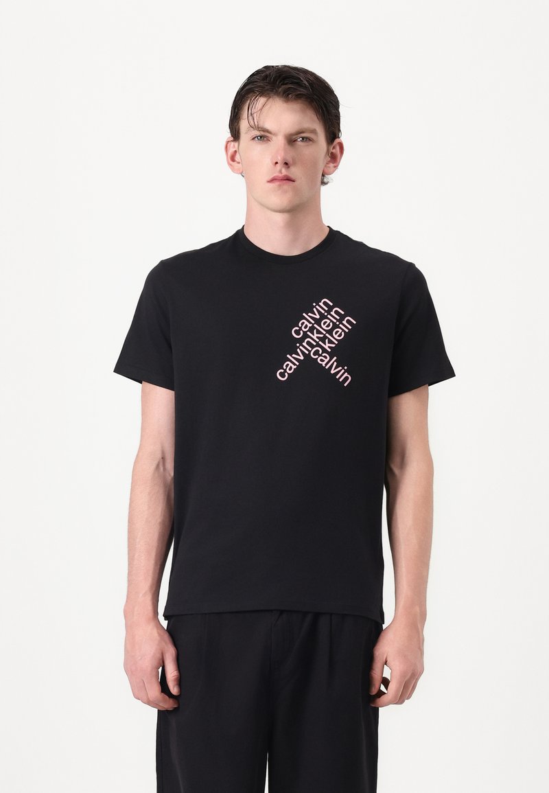 Zwarte katoenen t-shirt met een diagonaal, wit en roze herhalend tekstlogo in een vet sans-serif lettertype aan de linkerkant van de borst.