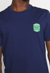 Navy blue cotton t-shirt with short sleeves, featuring a green and white embroidered Lacoste Club logo on the chest. Smooth texture.
