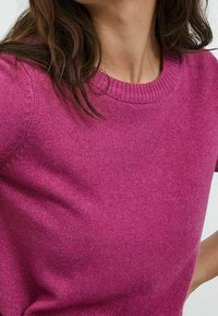 Femme portant un pull doux à manches courtes et col rond, de couleur magenta vif avec un tissu tricoté texturé visible.