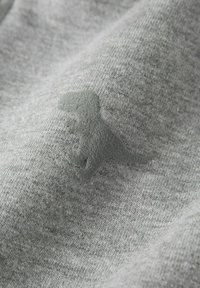 Sivi tkanina s mekanom teksturom sadrži suptilan, urezani logo dinosaura u nešto tamnijoj sivoj boji. Dizajn je jednostavan i minimalistički.