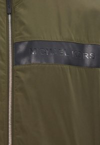 Maslinasto zelena najlon jakna sa crnim kožnim horizontalnim panelom sa urezanim "MICHAEL KORS" logom i srebrnim zip zatvaranjem.