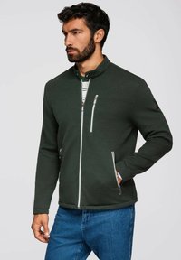 Mörkgrön zip-up jacka i strukturerat material, hög krage, två framfickor med dragkedja och vita dragkedjeaccenter. Bärs med blå jeans.