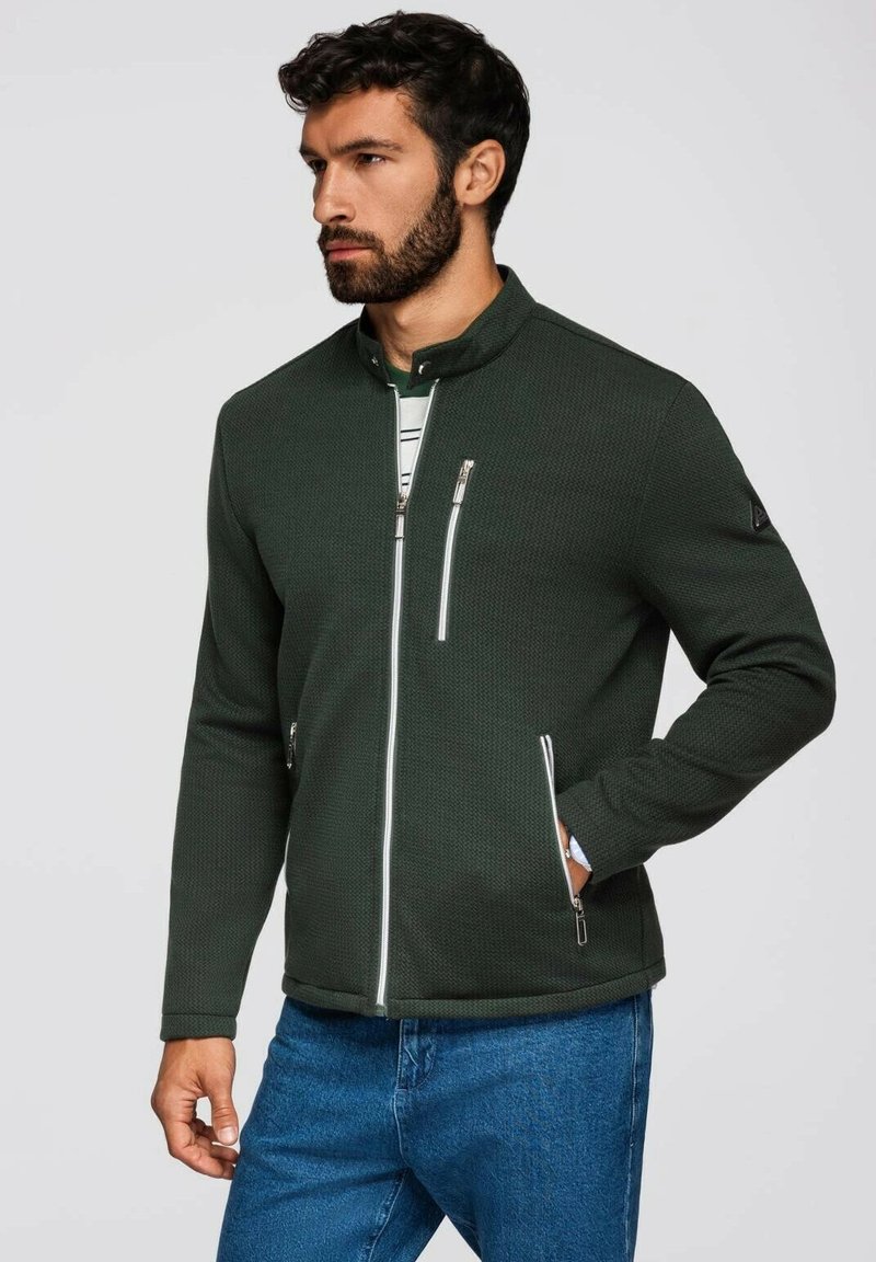 Mörkgrön zip-up jacka i strukturerat material, hög krage, två framfickor med dragkedja och vita dragkedjeaccenter. Bärs med blå jeans.