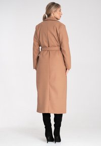 Długi, zapinany płaszcz z kamelowej mieszanki wełny, z wyłamiącym kołnierzem i dopasowaną sylwetką, zestawiony z wysokimi na kolana czarnymi zamszowymi botkami.