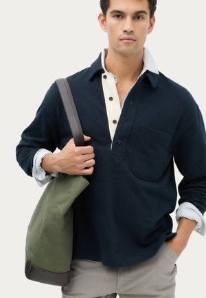 Junger Mann trägt einen marineblauen Pullover über einem gestreiften Hemd mit Kragen, hält eine olivgrüne Tragetasche mit braunem Riemen und steht vor einem hellen Hintergrund.