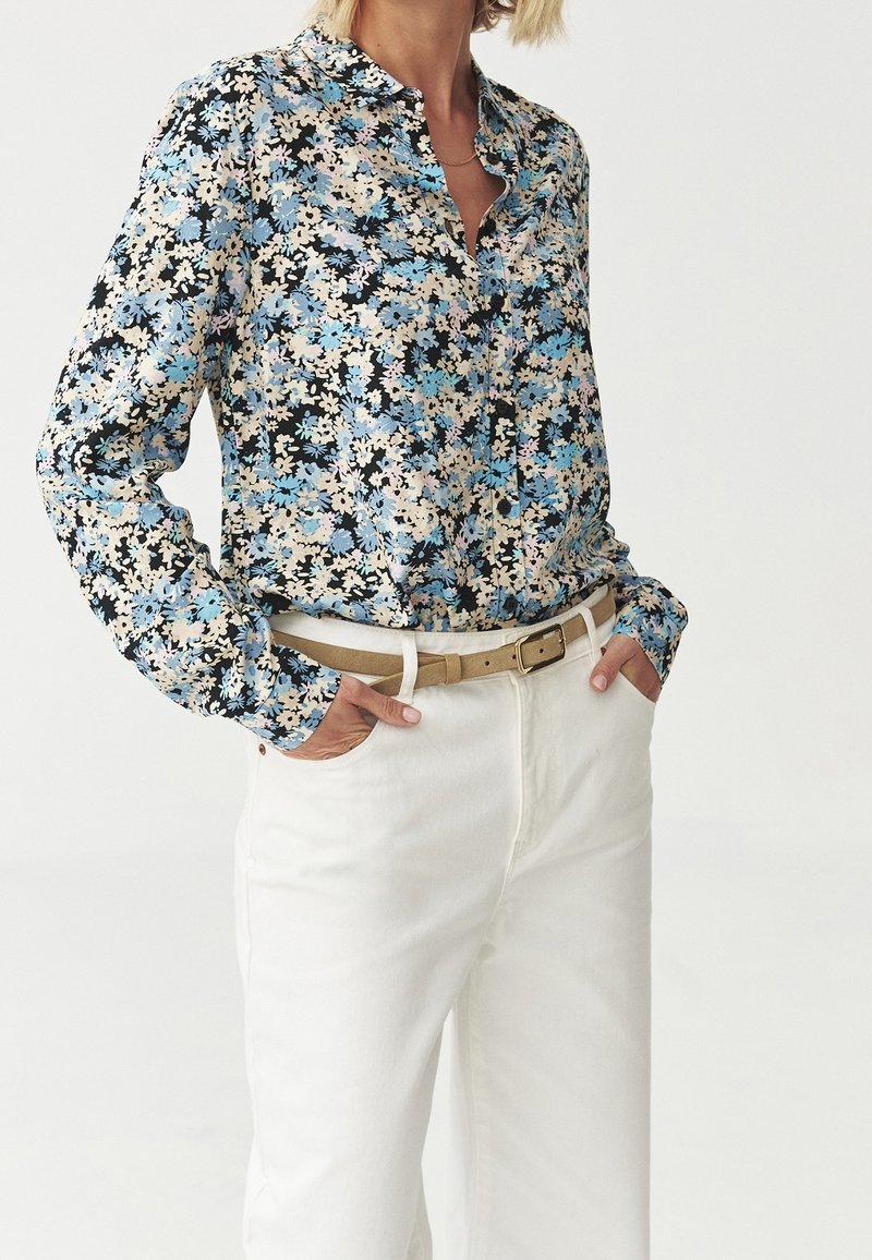 Květinová blůza s vícerozměrným vzorem v modré, růžové a černé barvě z lehkého materiálu, spojená s bílými kalhotami a béžovým pásem.