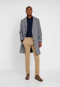 Płaszcz w wzór houndstooth w czerni i bieli, noszony na granatowej koszuli. Beżowe chinosy i brązowe buty dopełniają strój.