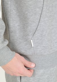 Harmaa huppari, jossa on pehmeä tekstuuri, ribatut hihansuut, sivutasku ja pieni merkkilappu sauman kohdalla. Lähikuva kankaasta.