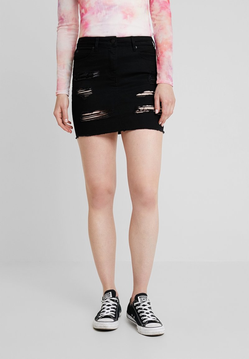 Personne portant une mini-jupe en denim noir déchiré, un haut à manches longues tie-dye rose et blanc, et des baskets Converse noires, debout sur un sol gris.