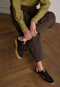 Black and burgundy sneakers with a textured upper, round toe, and gum sole. Grey knit sweater and brown pants. Natural wood floor.