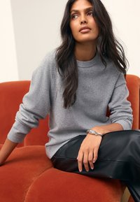Gray knitted sweater with a crew neckline, worn over black leather pants. The model's wrist features a silver bracelet, seated on a rust-colored couch.