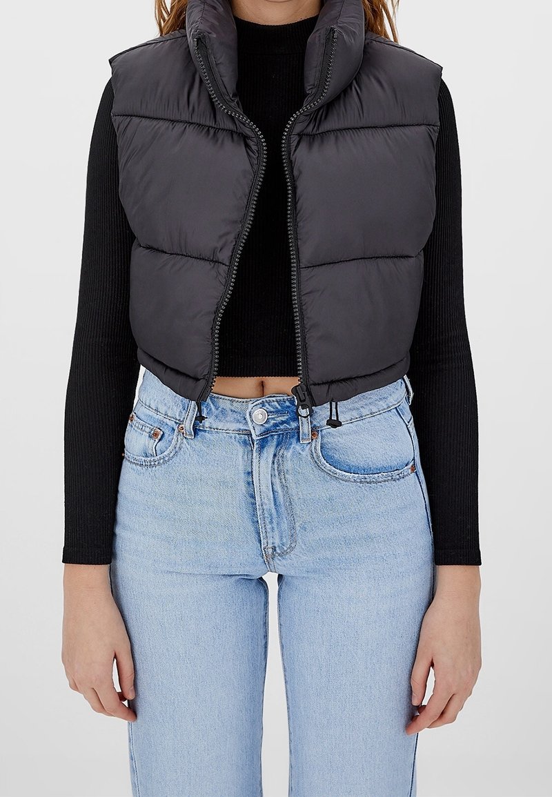 Woman wearing a black cropped puffer vest over a black long-sleeve top with light blue high-waisted jeans against a plain background.