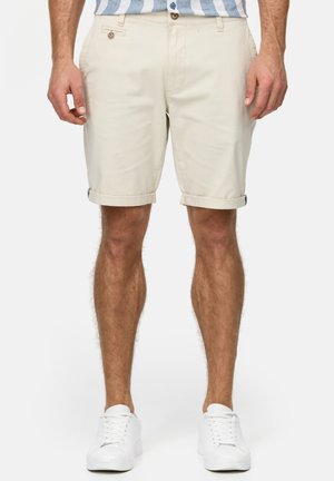 Beige bomuldsshorts med opsmøgede bukser, forlommer og knaplukning. Kombineret med hvide sneakers på en ensfarvet baggrund.
