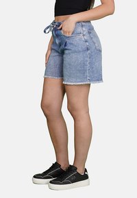 Shorts in denim azzurro chiaro con orlo sfrangiato, dotati di vita con lacci e tasche frontali, abbinati a sneakers nere.