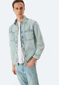 Vaaleansininen farkkutakki napittavalla edessä, kahdella rintataskulla ja rento istuvuus, yhdistettynä samanlaisiin vaaleansinisiin farkkuihin ja valkoiseen paitaan.