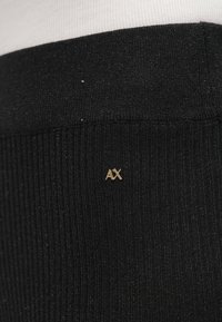 Tissu noir côtelé avec un petit logo "AX" doré en accent. La texture semble douce avec une légère brillance, adaptée pour une tenue décontractée.