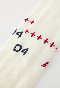 Valged kudumisokka, millel on tumesinised ja punased tikitud numbrid ja sümbolid. Mustris on "+" märgid ja peen ribakujundus.