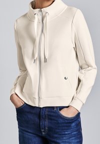 Chaqueta beige claro con cremallera, cuello alto, cordones ajustables y dos bolsillos delanteros. Tejido liso con detalles de costuras mínimas.