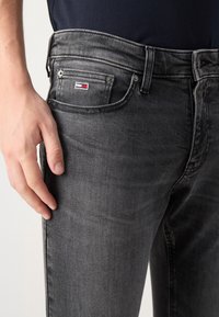 Dark gray denim jeans with a smooth texture, featuring a small embroidered logo, five-pocket design, and a straight fit.