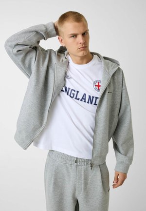 Junger Mann richtet seine kurzen Haare, trägt einen grauen Kapuzenpullover mit Reißverschluss, passende Jogginghose und ein weißes T-Shirt mit dem Schriftzug England und rotem Kreuz-Emblem.