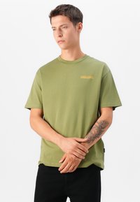 Olivza krāsas kokvilnas T-krekls ar īsām piedurknēm, apaļu kakla izgriezumu un mazu zeltītu logotipu kreisajā krūšu pusē. Vienkāršs dizains.