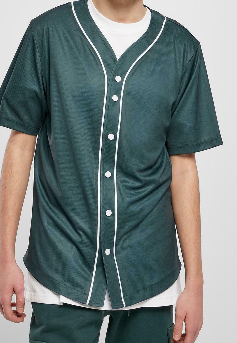 Homme portant un maillot de baseball vert foncé avec des boutons blancs et des bordures sur une chemise blanche, assorti d'un pantalon assorti.