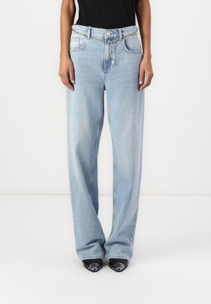 Persoon die lichtblauwe wijd uitlopende spijkerbroek draagt met een zilveren kettingriem en zwarte schoenen met een puntige neus, staand tegen een effen witte achtergrond.