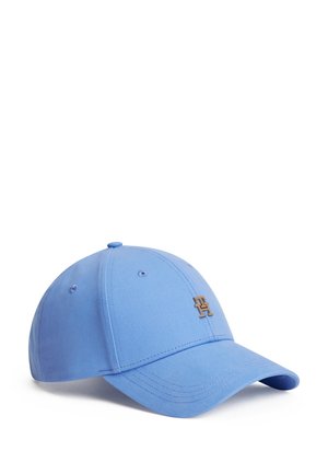 Femme portant une casquette de baseball bleu clair et un t-shirt blanc avec des fleurs brodées colorées sur la poitrine, ajustant sa casquette avec la main.