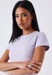 Camiseta ajustada de color lavanda con mangas cortas y cuello redondo; presenta un logo bordado en blanco en el pecho; textura de tela suave.