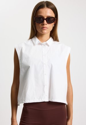 Biały, bezrękawnik skrócony z zapięciem na guziki z przodu. Gładka tekstura, koszulowy kołnierzyk. W połączeniu z brązowymi legginsami z wysokim stanem.